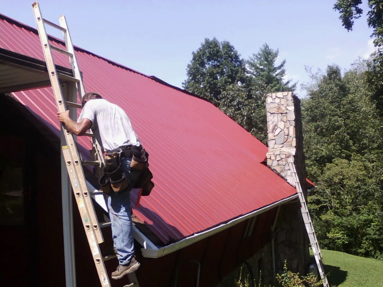 Roofer on a ladder beside a red metal roof and stone chimney — Soffit & Fascia Repair by Roofing Brothers in Lincoln University